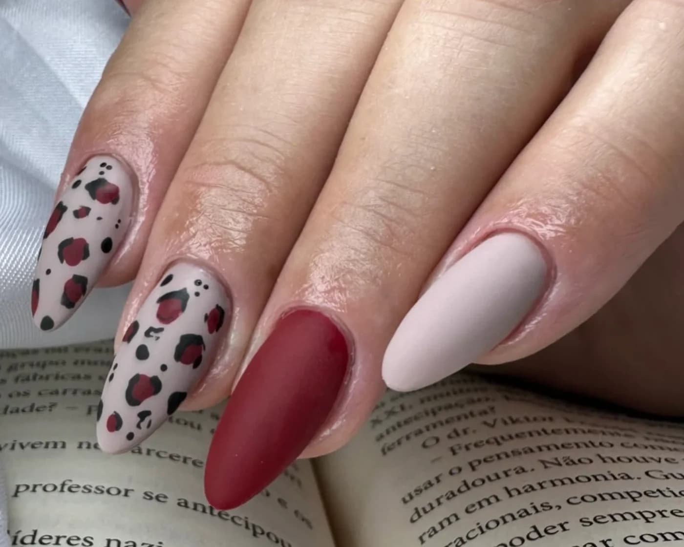 Unhas amendoadas em nude e vinho fosco, com estampa de oncinha em preto e vinho, apoiadas sobre um livro aberto.