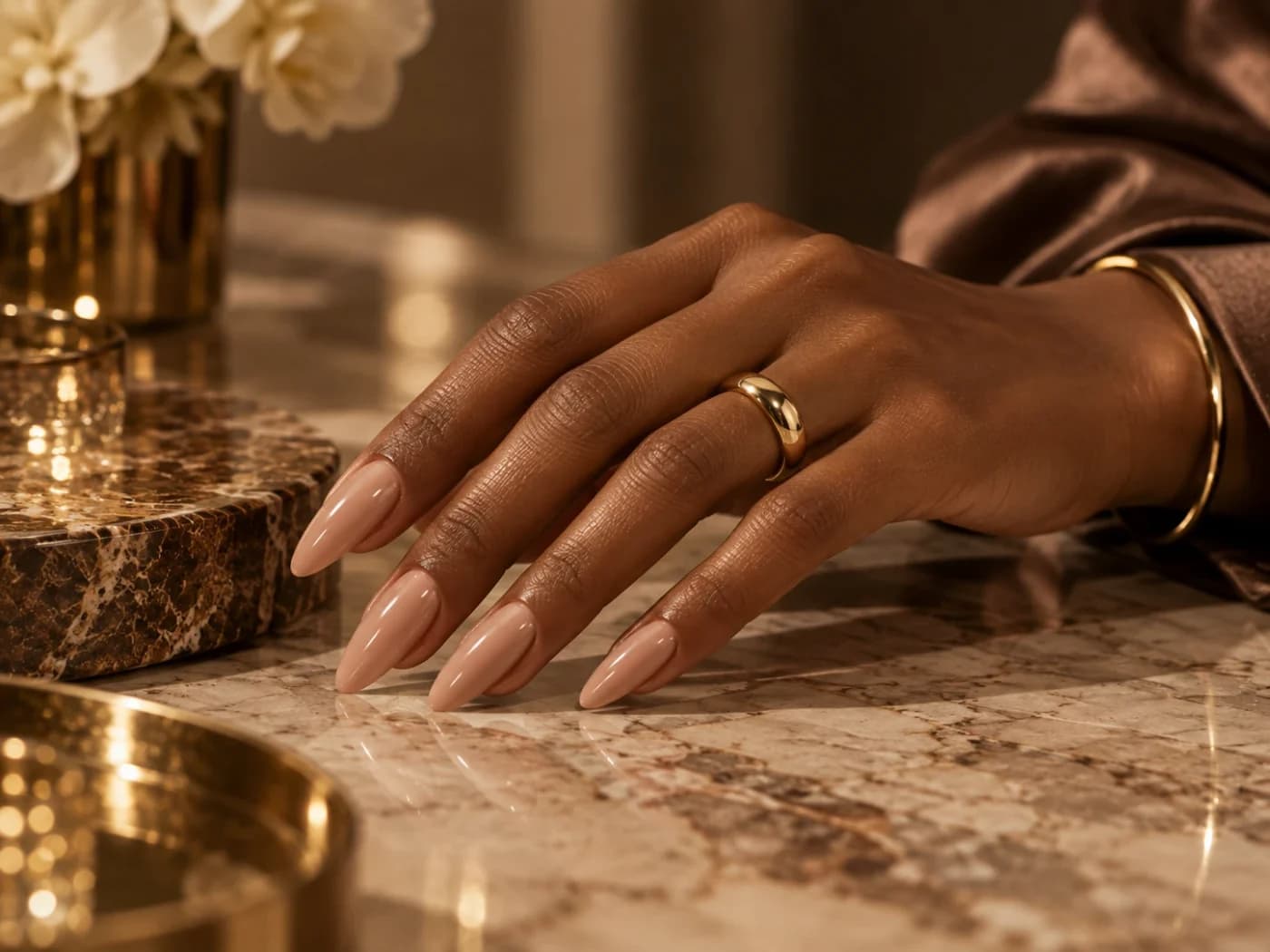 Mão com unhas alongadas em formato amendoado e acabamento nude acetinado, apoiada em bancada de mármore.